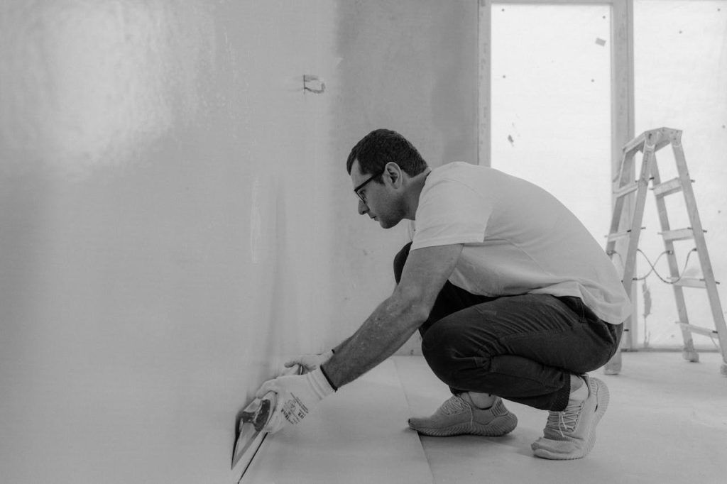 grayscale-photo-of-a-man-painting-a-house-6474344 Monochrome image of a man renovating an interior wall using plaster tools.
